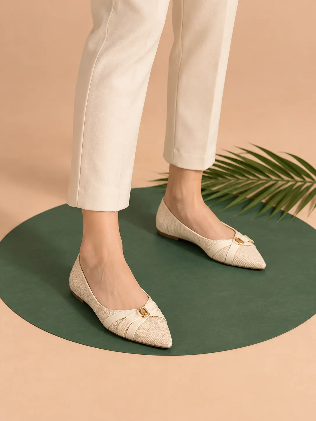 Model wearing elegant women’s flat shoes while standing on an emerald green oval over a warm peach studio floor with tropical palm styling.