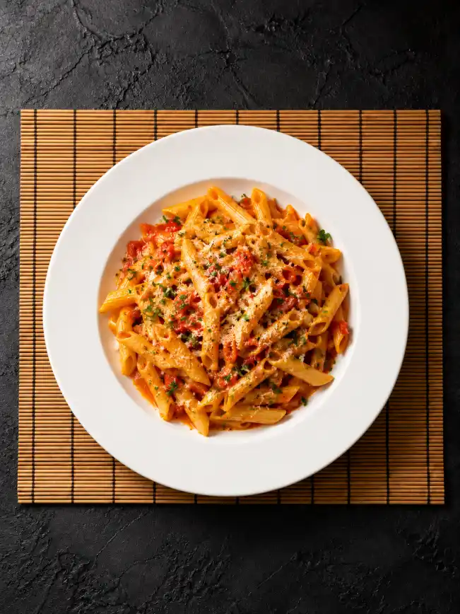 Overhead creamy tomato penne pasta on a white ceramic plate placed on a bamboo mat over a dark textured background.