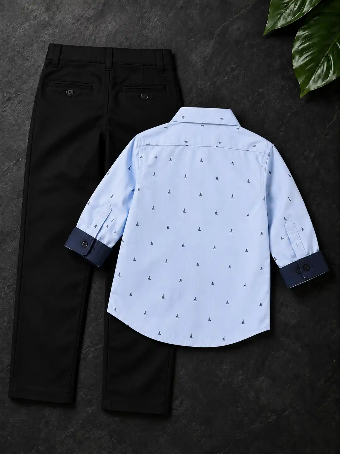 Back view clothing flat lay on a dark charcoal surface with black trousers, tropical leaves, soft shadows, and premium fashion styling.