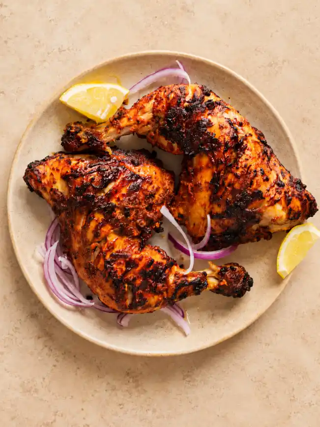 Overhead view of charred tandoori chicken served with red onion slices and lemon wedges on a warm beige ceramic plate.