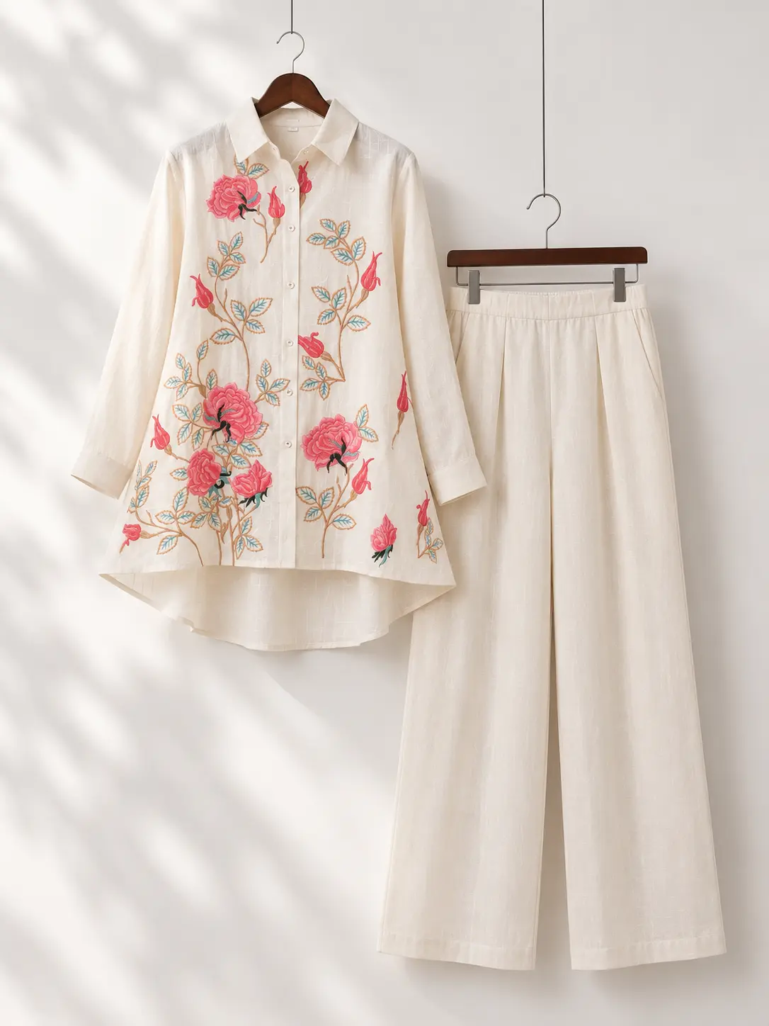 Cream floral embroidered long shirt with matching wide-leg pants hanging on wooden hangers against a minimal white studio background.