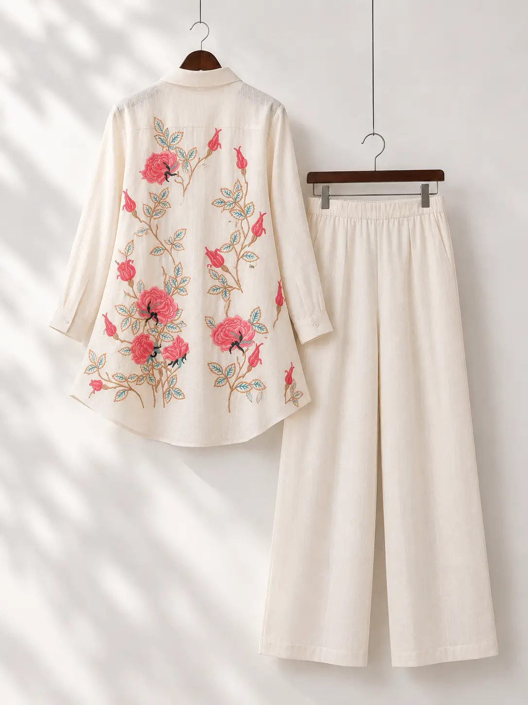Premium back-view product photo of a cream floral embroidered kurti and matching pants hanging on wooden hangers in a minimal studio.
