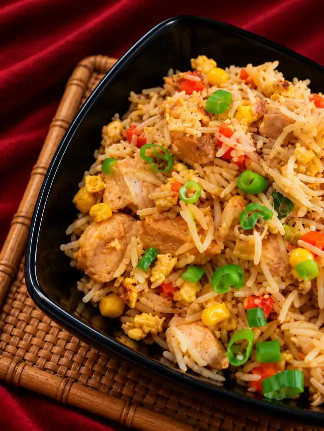 Extreme close-up of chicken fried rice with chicken, egg, corn, peppers and spring onions in a glossy black triangular bowl.