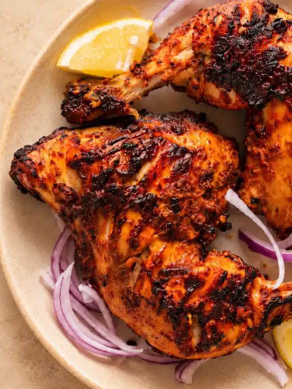 Extreme close-up of charred tandoori chicken with red onion slices and lemon wedges on a warm beige ceramic plate.