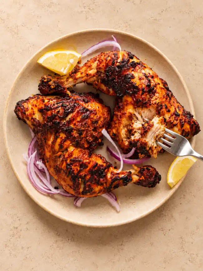 Overhead view of charred tandoori chicken with lemon and red onion on a warm beige ceramic plate as a fork lifts a small juicy bite.