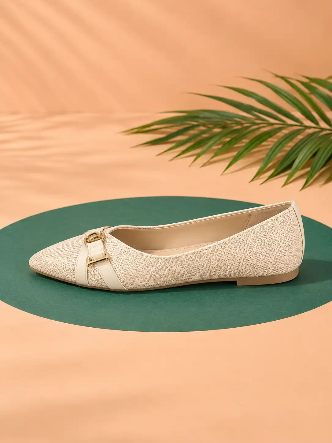 Elegant women’s pointed flat shoe in side view placed on an emerald green oval over a warm peach studio surface with palm leaves.