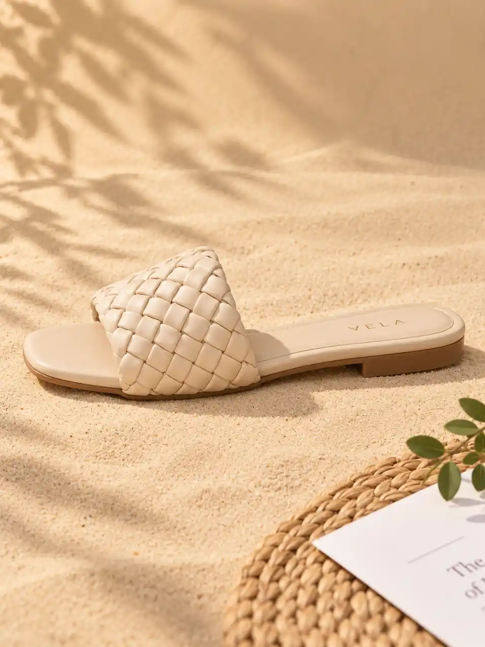 Single women’s flat sandal shown in side view on warm beige sand with woven mat, white paper and subtle green botanical styling.