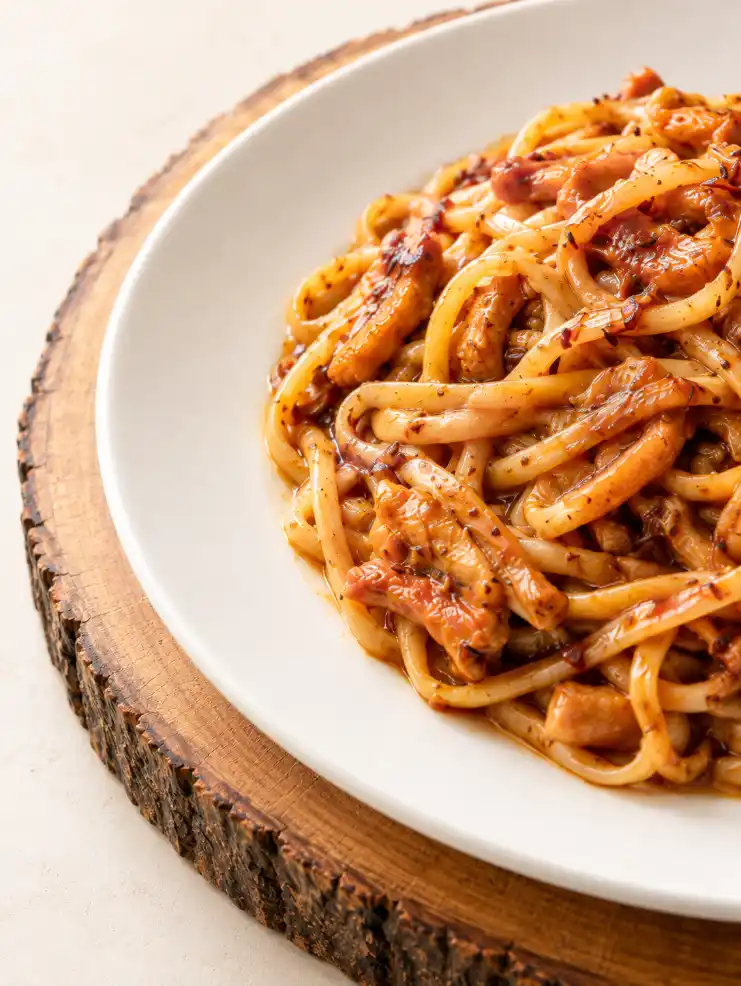 Close-up of glossy stir-fried noodles with meat on a white ceramic plate over a rustic wooden slab.