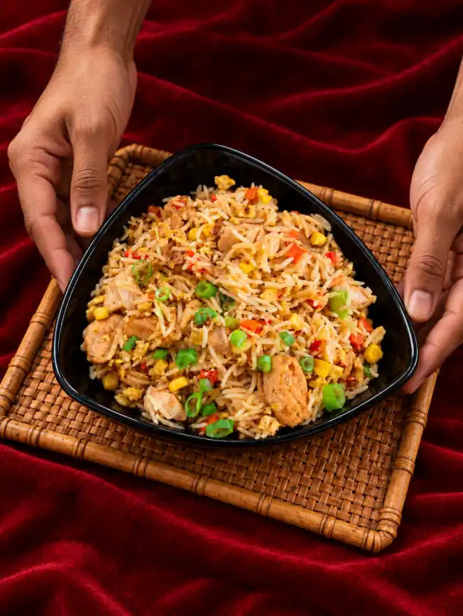 Chef hands placing a black triangular bowl filled with chicken fried rice on a woven bamboo mat over a deep red fabric background.