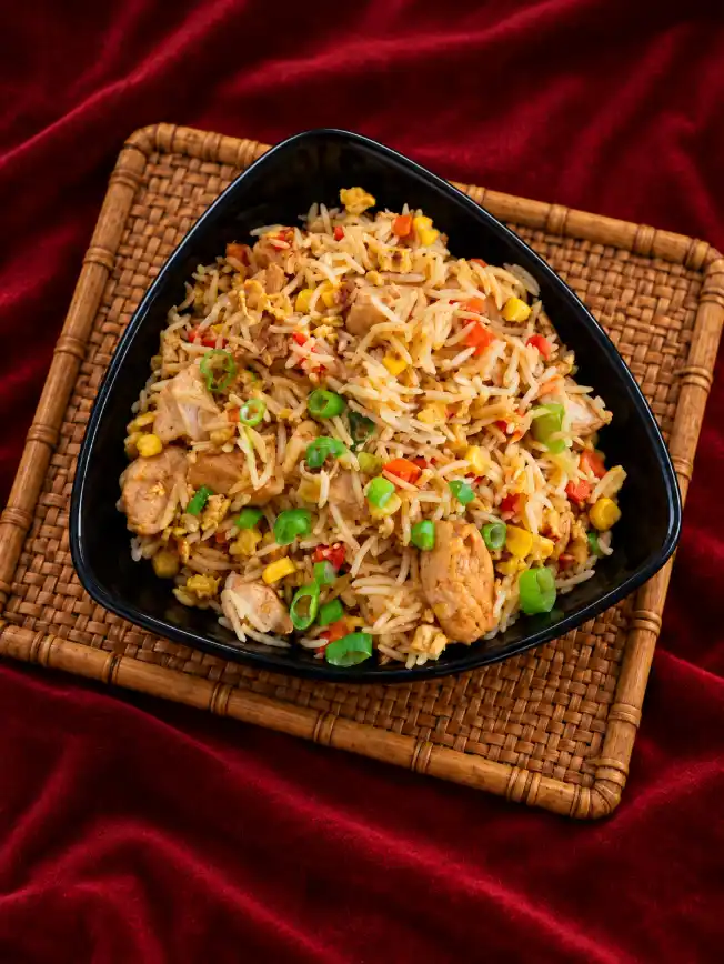 Chicken fried rice with vegetables and chicken pieces served in a black triangular bowl on a bamboo mat over a deep red background.