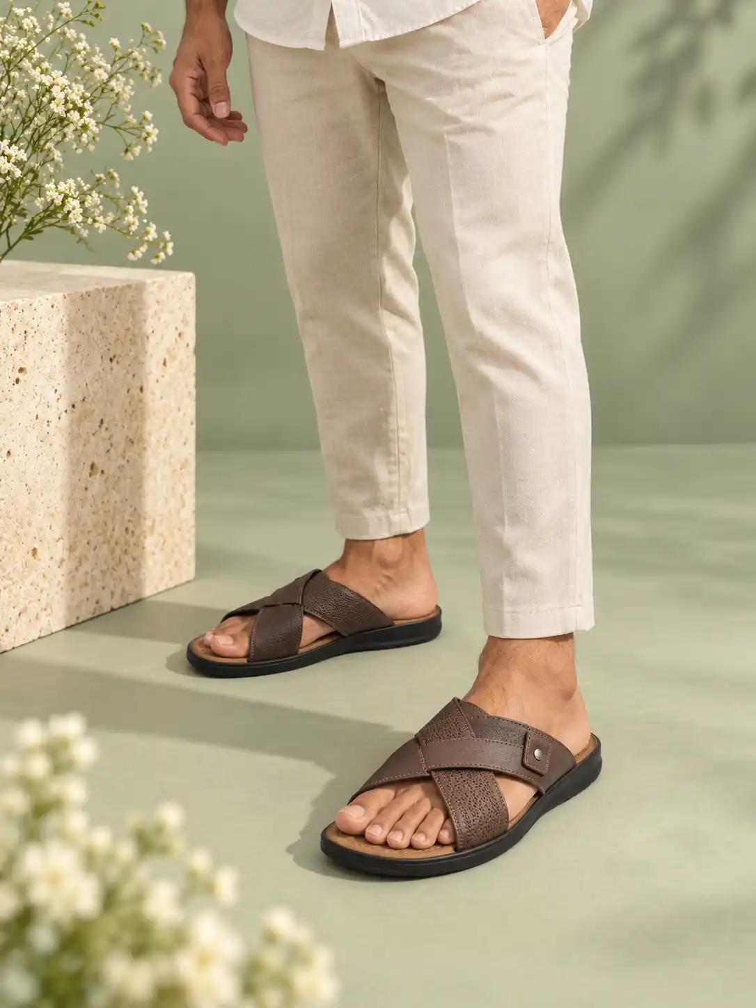 Fashion model wearing footwear in a soft sage green studio with a cream stone pedestal, delicate white flowers, and warm natural lighting.